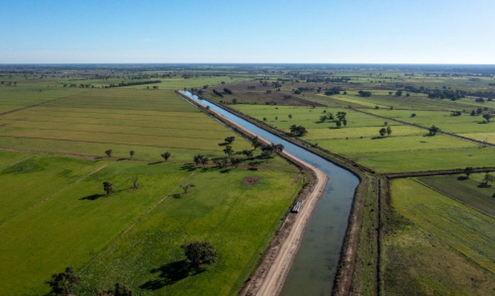Aerial drone shot of a large irrigation channel filled with water and running between vast green paddocks. A bright blue sky is in the background. 