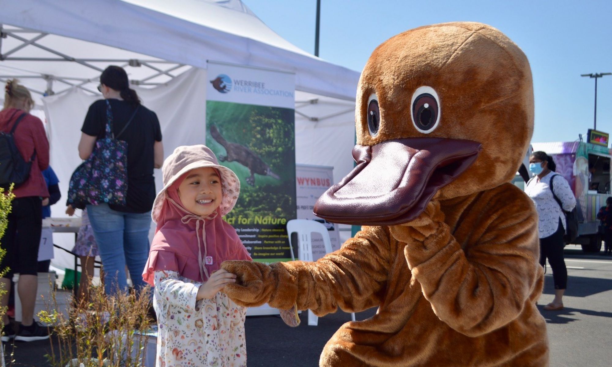 A young child at a fair, holding the hand of an adult sized costumed platypus. The platypus is kneeling down. 