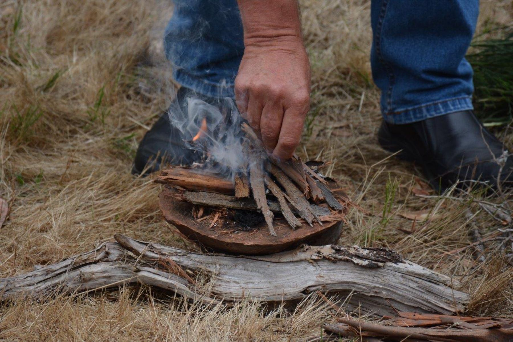 Image of a person hands and feet. They are bent down towards a small smoking ceremony fire, bark in a dish leaning on a large piece of wood. 