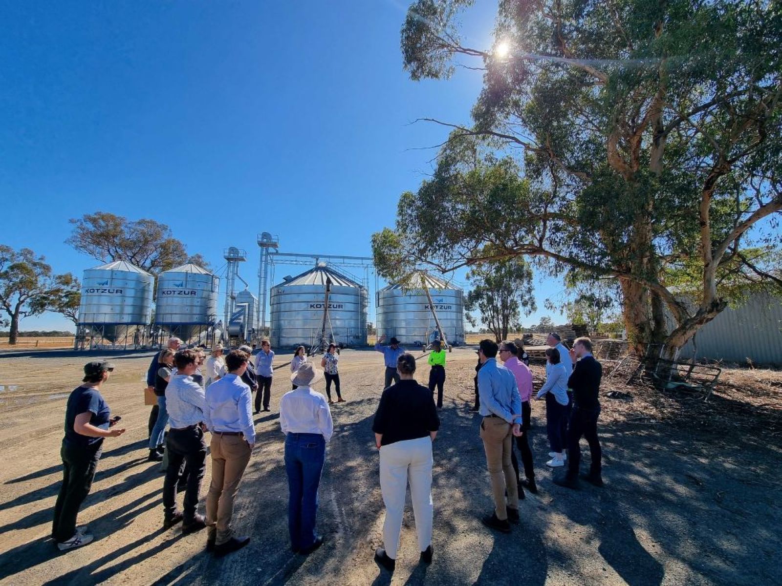 IDC members and two farmers standing in a circle in the shade of a large gum tree. In the background are four steel silos