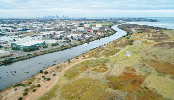 Kororoit Creek Altona. Aerial view of a coastal landscape featuring a winding creek bordered by industrial buildings on one side and expansive wetlands on the other. The wetlands display patches of green, brown, and rust‑coloured vegetation with walking tracks running through them. The creek flows toward a bay in the distance. A large suburban and industrial area stretches toward the horizon, with a city skyline visible far in the background under a cloudy sky