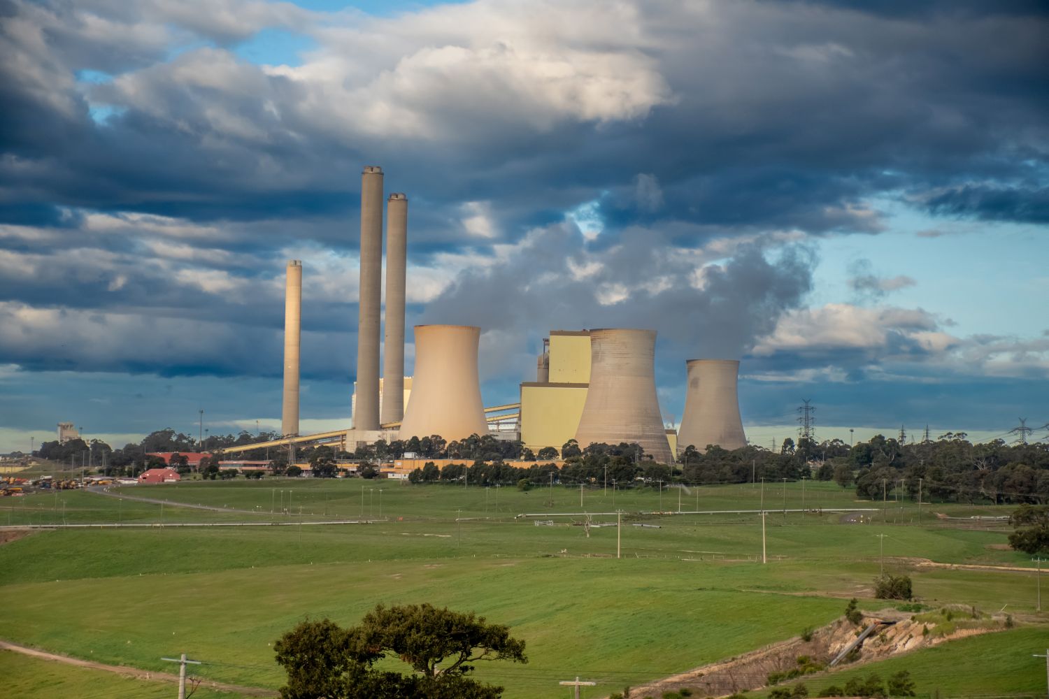 A power plant with cooling towers and smokestacks in the background with lush fields in the foreground  