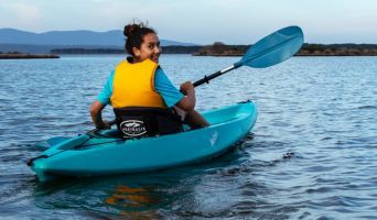 a young woman in a lifejacket kayaks down a river and turns to the camera.