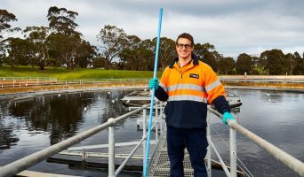 A young man in orange hi-viz and safety goggles stands atop a recycled water tank.